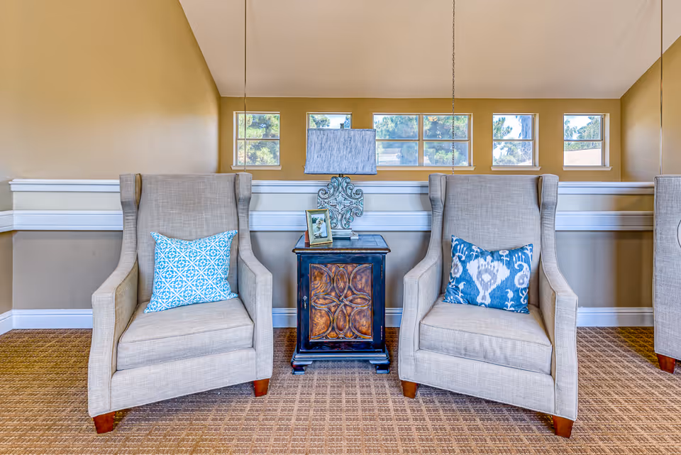 Two upholstered armchairs with patterned throw pillows flank a decorative side table and lamp in a bright seating area.