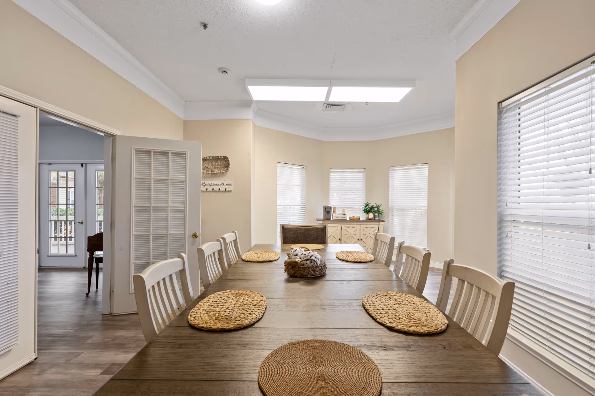 Long communal dining room with a wooden table set with woven placemats, white chairs, and windows with blinds.
