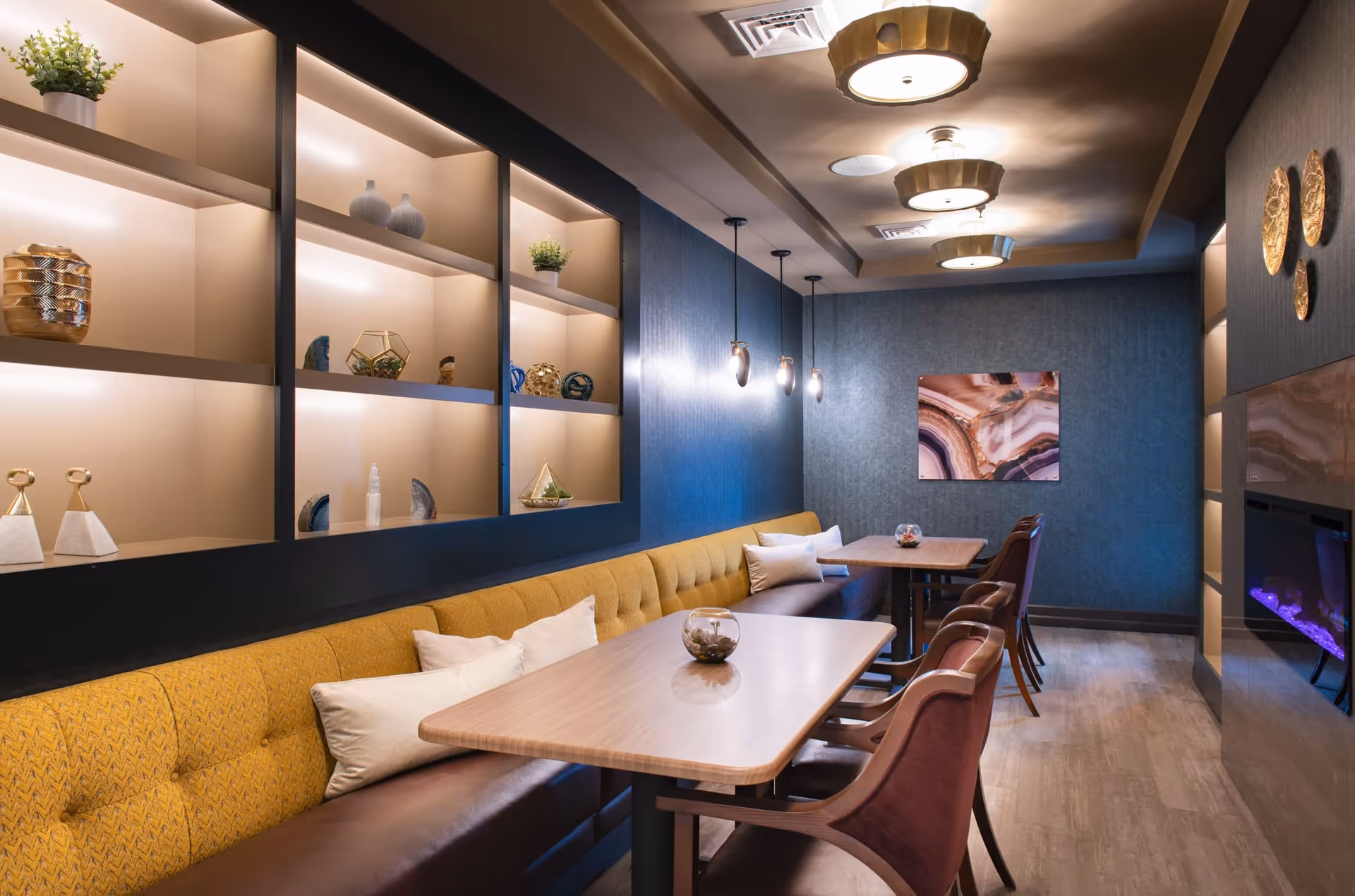 A cozy dining area in a senior living facility featuring two wooden tables with cushioned chairs and a long mustard-yellow upholstered bench with pillows. The walls are decorated with built-in shelves displaying decorative items, modern pendant lights, and a piece of abstract wall art. There is also a modern electric fireplace on the right side.