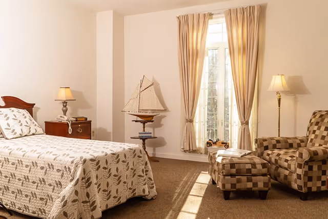 A cozy bedroom in an assisted living facility featuring a single bed with a leaf-patterned quilt, a wooden nightstand with a lamp and telephone, a decorative model sailboat on a small round table, a window with beige curtains letting in natural light, and a checkered armchair with a matching ottoman next to a floor lamp.