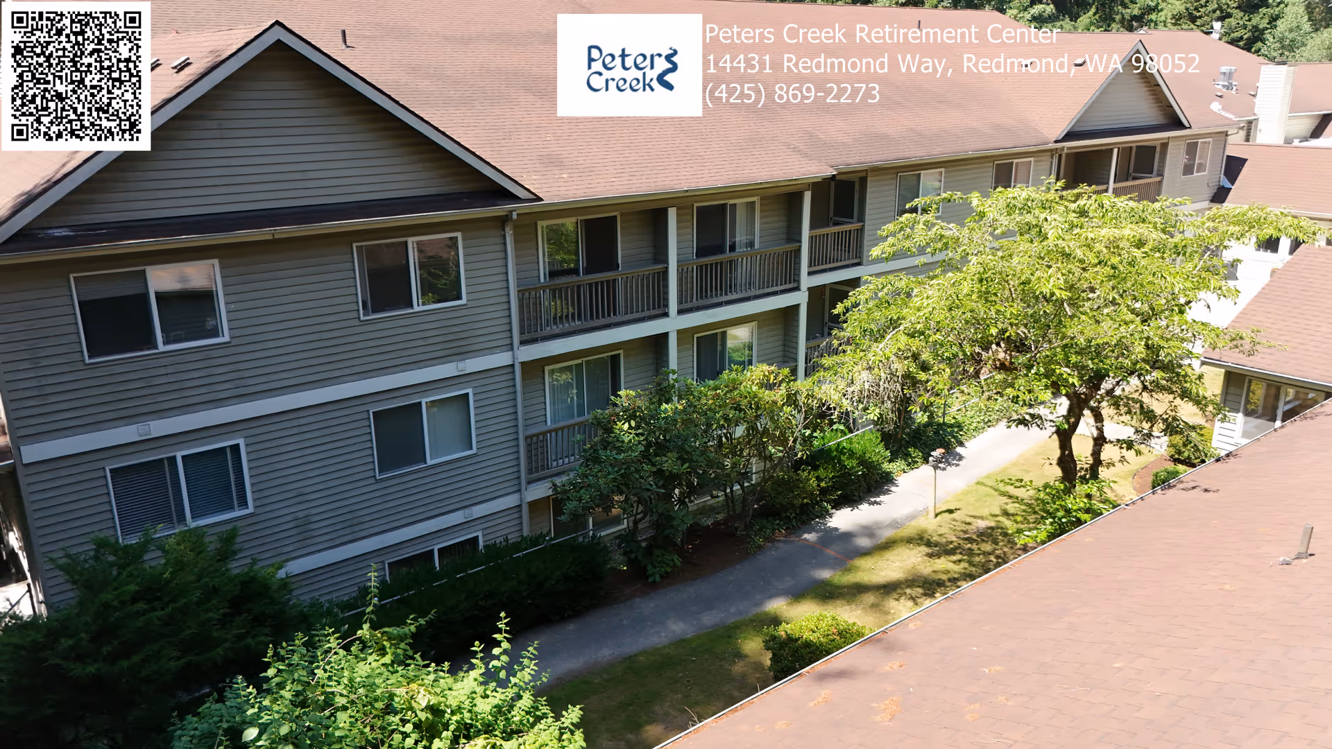Exterior view of a multi-story residential building with beige siding and brown roof, surrounded by greenery including bushes and trees, with a paved walkway running alongside the building under a sunny sky.