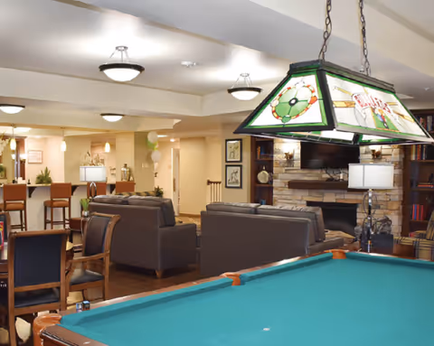 A bright common lounge with a pool table in the foreground, sofas around a stone fireplace, and a small dining/bar area behind.