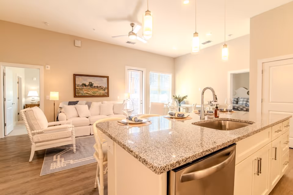 Bright and spacious open-concept living area featuring a large granite kitchen island with a sink and dishwasher, set with plates and wine glasses. The living room includes a white sofa, cushioned armchair, floor lamp, and a framed landscape painting on the wall. Two doorways lead to bedrooms, one with a visible bed and plaid bedding. The room is well-lit with pendant lights, ceiling fan, and natural light from windows and a glass door.