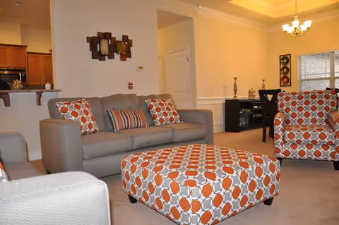 A cozy living room with a gray sofa adorned with patterned orange and white pillows, a matching patterned ottoman in the center, and an armchair with the same pattern. The room has beige walls, carpeted floor, and a dining area visible in the background with a dark wood table and chairs. There is a decorative wall piece above the sofa and a chandelier hanging from the ceiling.