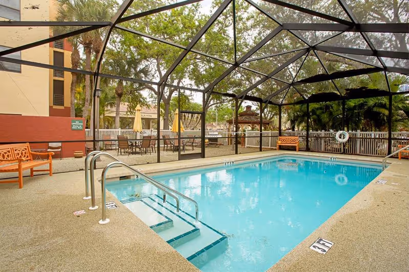 Screened outdoor swimming pool with steps, metal handrails, benches, patio tables and a gazebo beside the facility building.