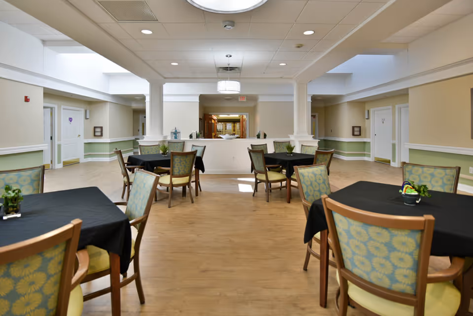 A spacious dining room with several tables covered in black tablecloths and surrounded by wooden chairs with patterned cushions. The room has light-colored walls with green wainscoting, a wooden floor, and a white ceiling with recessed lighting. There are small plants on the tables and a decorative light fixture hanging from the ceiling.