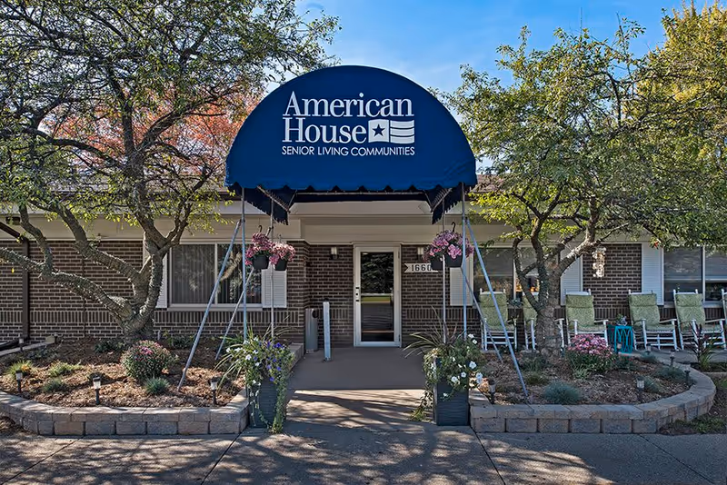 Entrance to American House senior living community with a blue awning displaying the name and logo. The entrance is flanked by landscaped garden beds with trees, flowers, and shrubs. Several green rocking chairs are visible on the right side near the building windows.