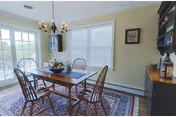 A dining room with a wooden table and six chairs placed on a patterned rug. The room has large windows with white blinds, a chandelier hanging above the table, and a cabinet with decorative items on the right side.