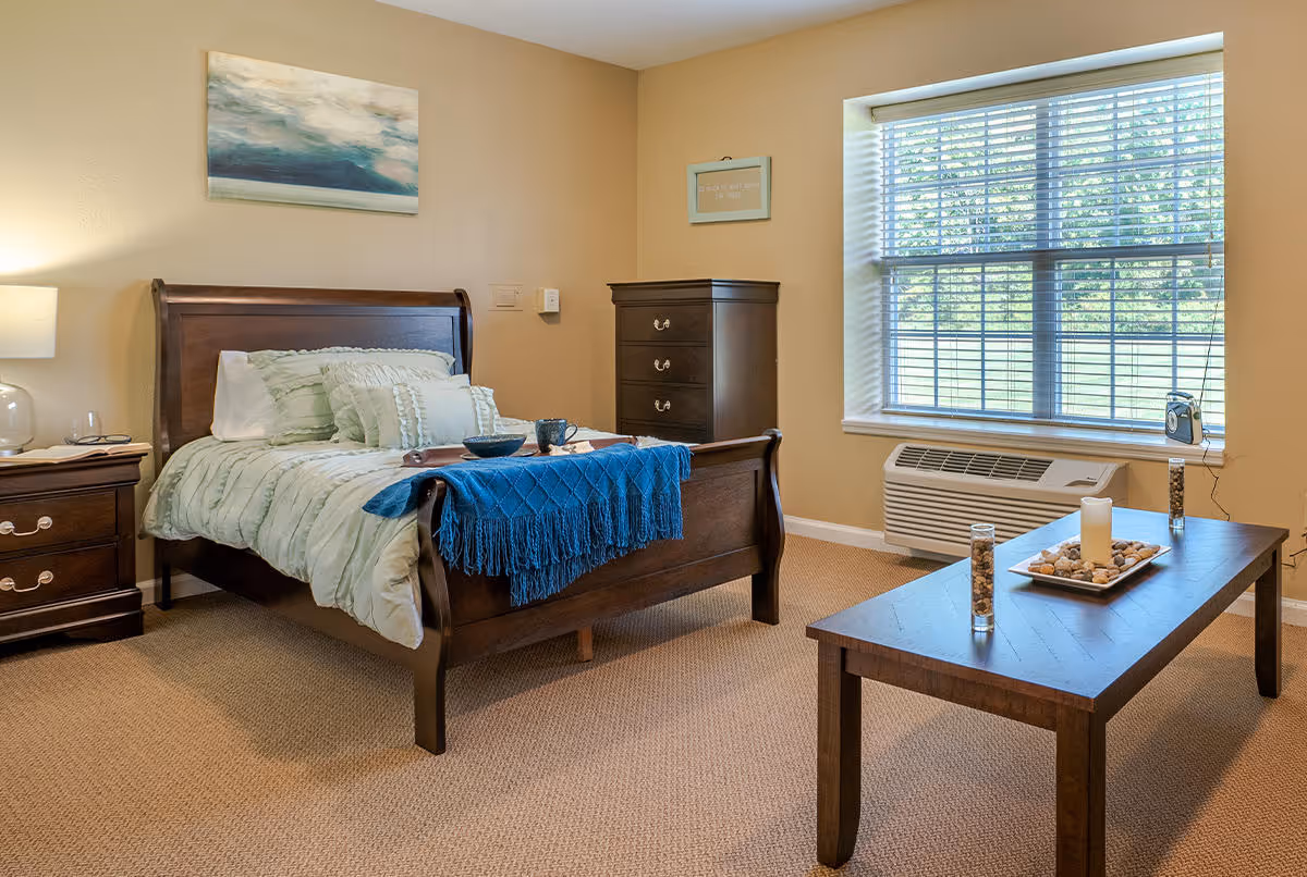 A cozy bedroom in a senior living facility featuring a wooden bed with light green bedding and a blue throw blanket. There is a wooden nightstand with a lamp and a glass, a tall wooden dresser, a large window with blinds, and a wooden coffee table with decorative items including a candle and vases filled with stones.
