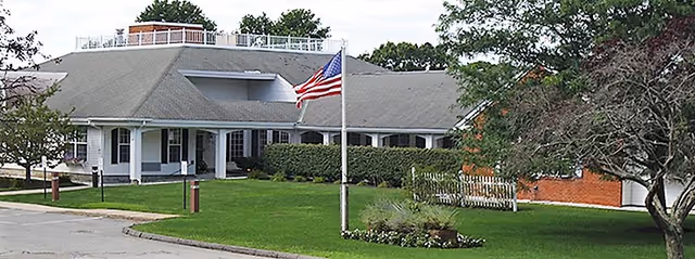 Exterior view of a single-story building with a gray roof and white pillars at the entrance. There is a flagpole with an American flag in front of the building, surrounded by green grass, shrubs, and trees under a partly cloudy sky.