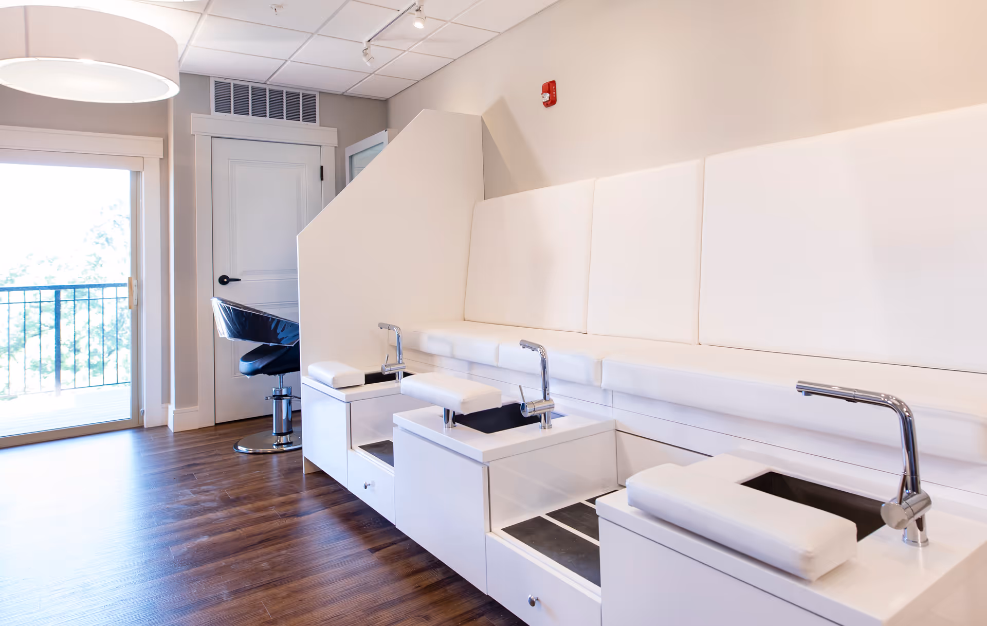 A bright and clean pedicure station in a salon area with white cushioned seating, foot baths with faucets, and a black salon chair near a closed white door. The room has wooden flooring and a large window with a view of greenery outside.