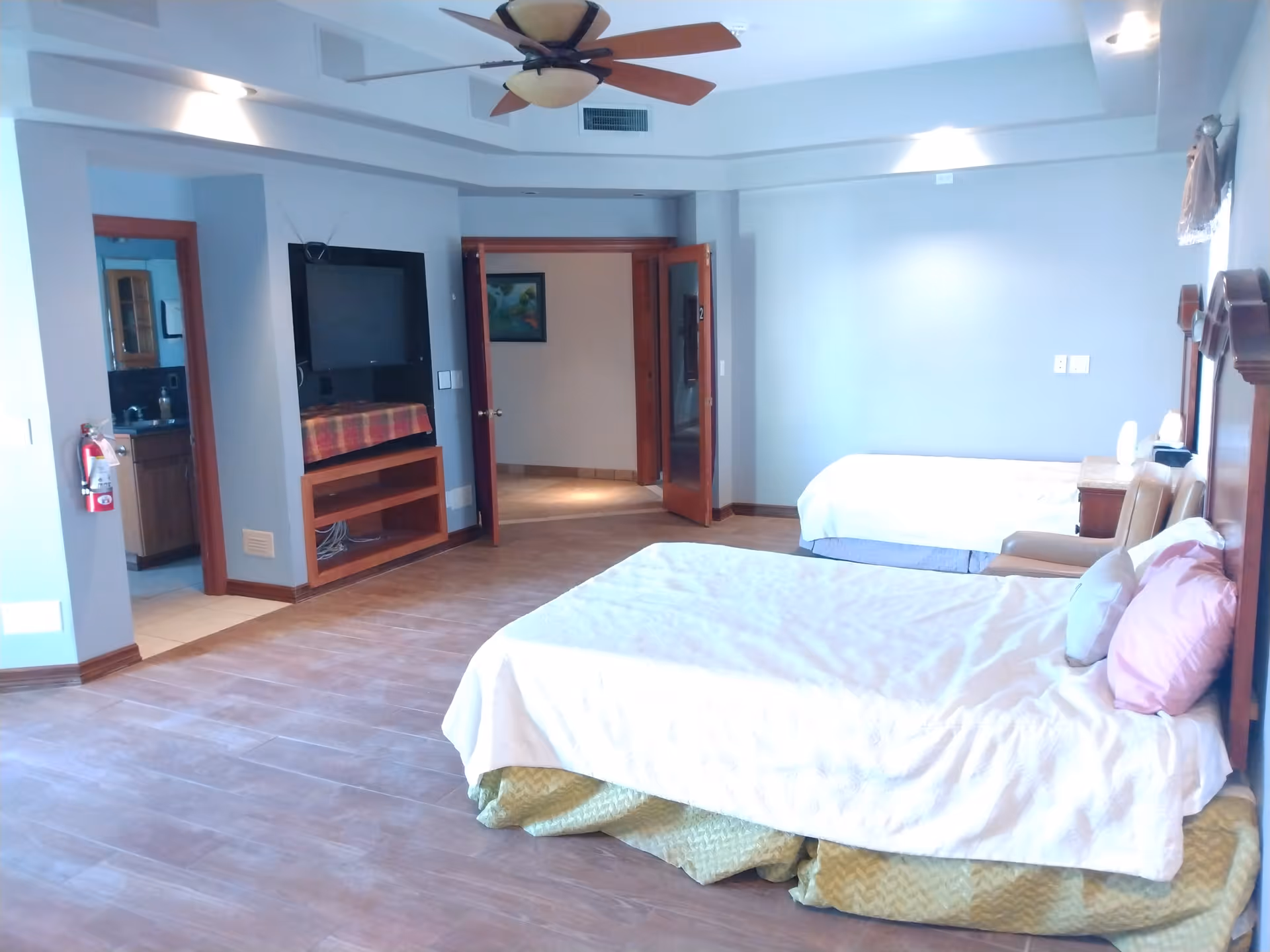 A spacious bedroom with two beds, a ceiling fan, a wall-mounted TV, and an open door leading to another room. The room has light blue walls, wooden trim, and a tiled floor. There is a small seating area with a chair next to one of the beds.