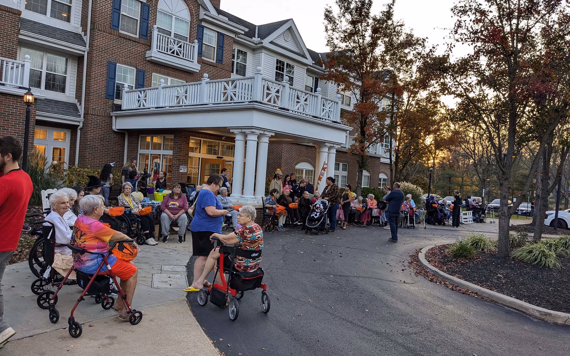 A crowd of residents and visitors gathered outside the front entrance of a multi-story brick senior living building at dusk.