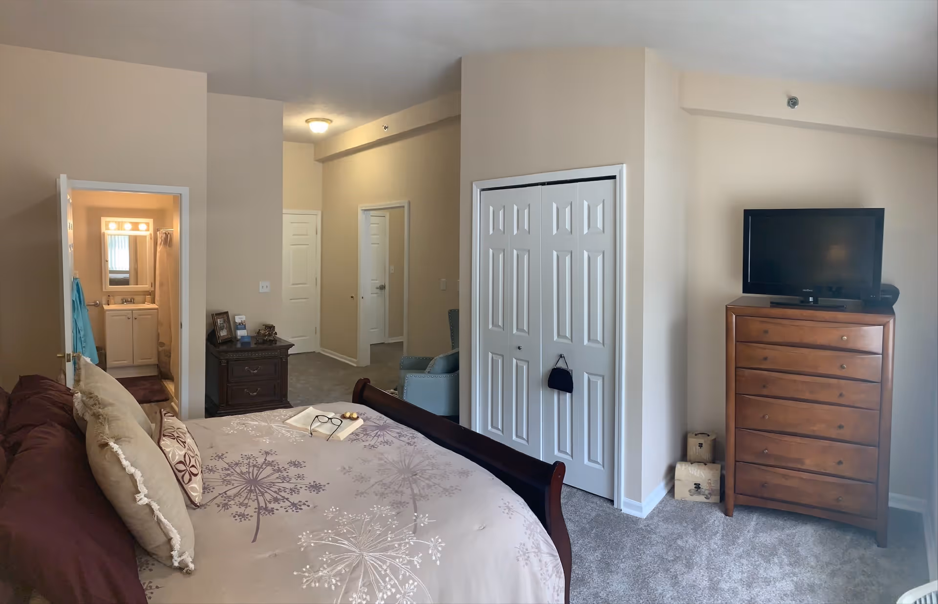 A bedroom in an assisted living facility featuring a bed with floral-patterned bedding and multiple pillows. To the left, there is an open door leading to a bathroom with a sink, mirror, and shower curtain visible. In the center, there is a small dark wooden nightstand with picture frames and decorative items. To the right, there is a white double-door closet and a wooden dresser with a flat-screen TV on top. The room has beige walls and carpeted floors.