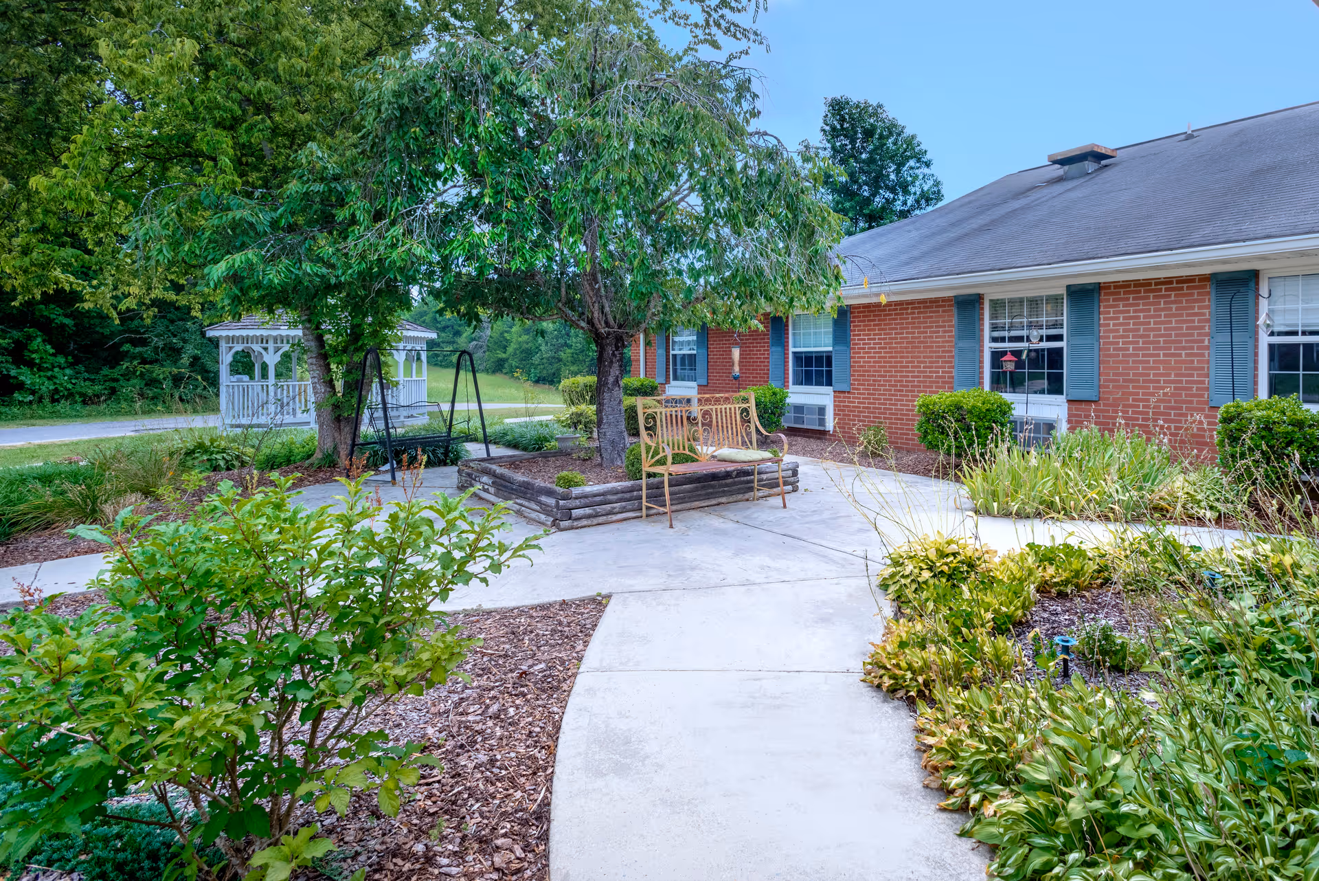 Outdoor garden area at The Courtyard at Deer Ridge featuring a paved walkway, green shrubs, a tree with a bench underneath, a swing, and a white gazebo in the background next to a red brick building with blue shutters and windows.