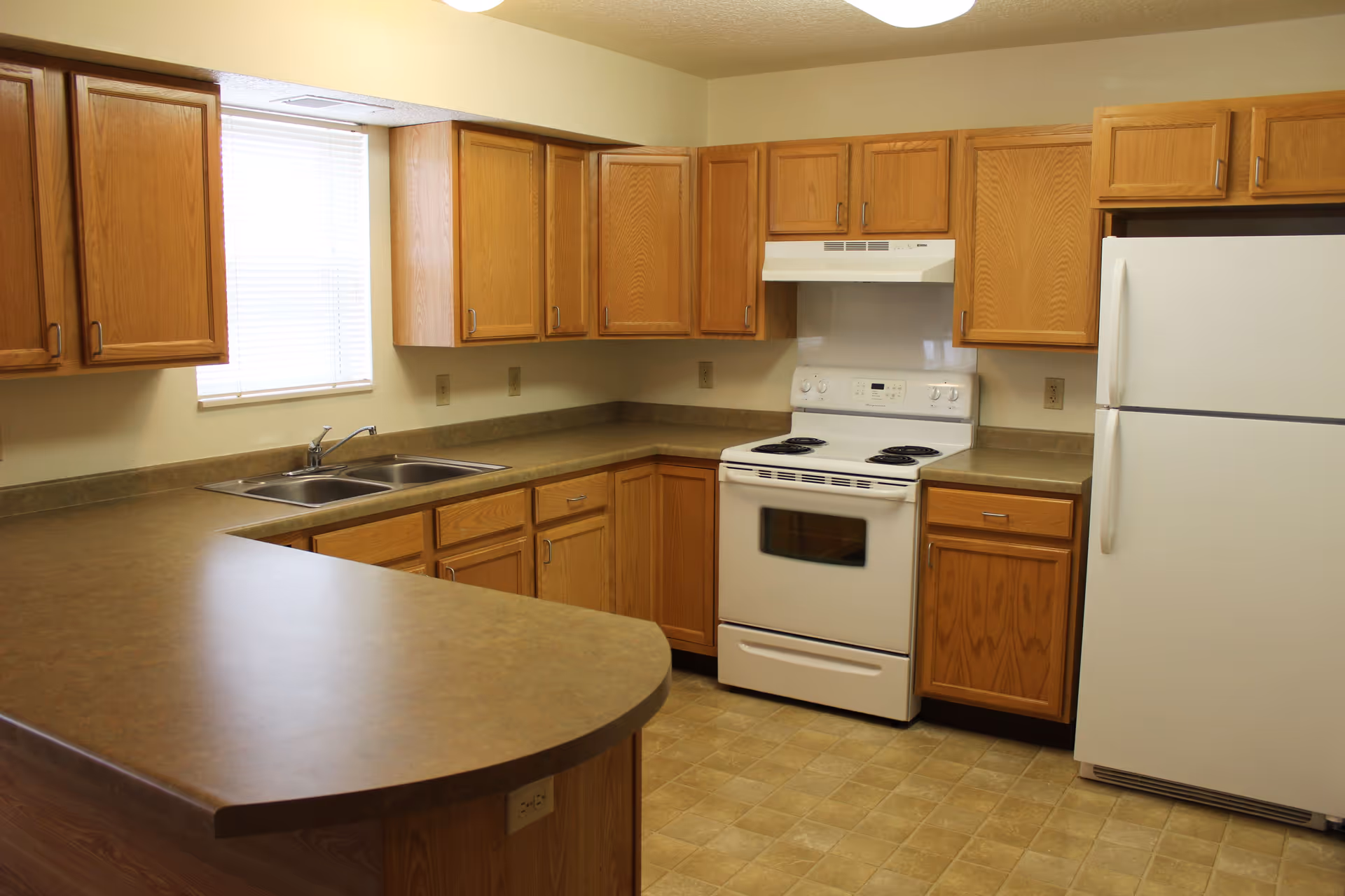 A kitchen with wooden cabinets, a white electric stove with oven, a white refrigerator, a double sink under a window with blinds, and a brown countertop with an extended breakfast bar.