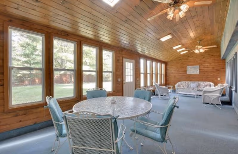 A spacious sunroom with wooden paneled walls and ceiling, featuring multiple large windows letting in natural light. The room is furnished with a round white metal table surrounded by six cushioned chairs, and a seating area with a floral-patterned sofa and two armchairs. Ceiling fans with lights are mounted on the ceiling.