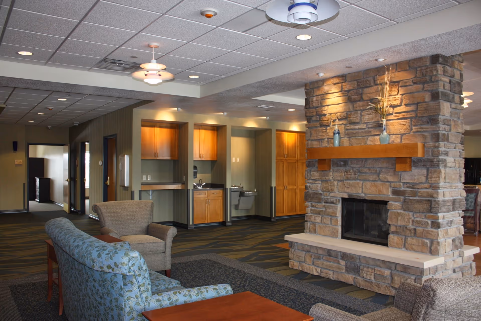 A cozy common area in a senior living facility featuring a stone fireplace with a wooden mantel decorated with vases and dried plants. The room has comfortable seating including a floral-patterned sofa and an armchair, with wooden tables. In the background, there are wooden cabinets, a drinking fountain, and a small sink area under warm lighting. The ceiling has recessed lights and hanging fixtures.
