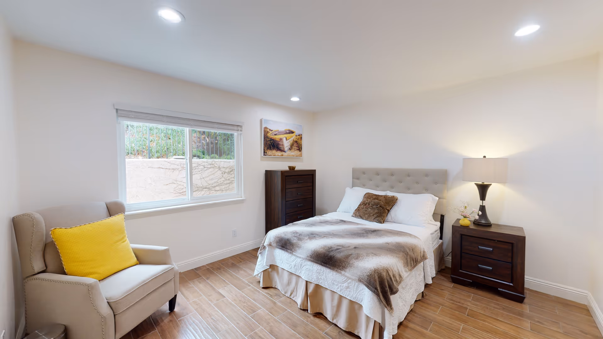 A cozy bedroom with a double bed featuring white and brown bedding, a tufted headboard, a wooden nightstand with a lamp and decorative items, a tall wooden dresser, a beige armchair with a bright yellow pillow, and a window with a view of an outdoor wall and greenery.