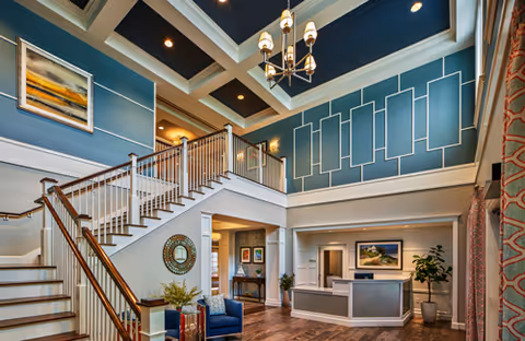 Bright two-story lobby with a central staircase, reception desk, seating area, and decorative blue walls and chandelier.
