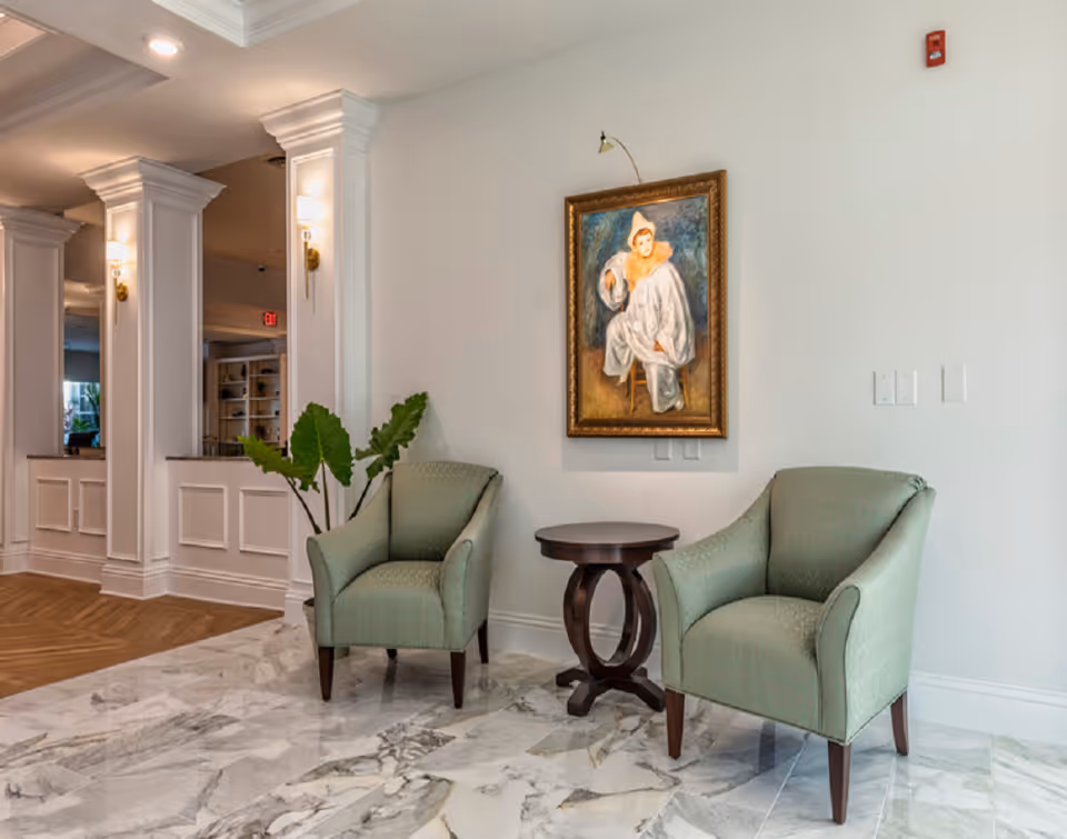 Two green armchairs flanking a small round table in a bright lobby with marble flooring, framed painting, and decorative columns.