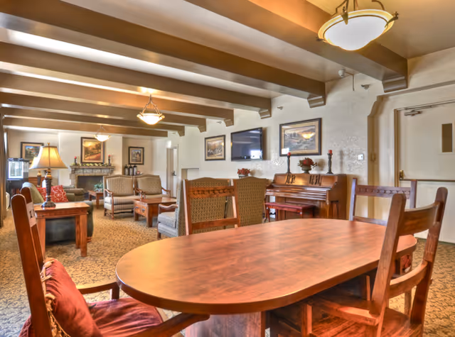 A cozy living room area in California Mission Inn featuring wooden furniture including a large oval table with chairs, a piano against the wall, several armchairs, a fireplace, framed artwork, and warm lighting from ceiling fixtures and a table lamp.