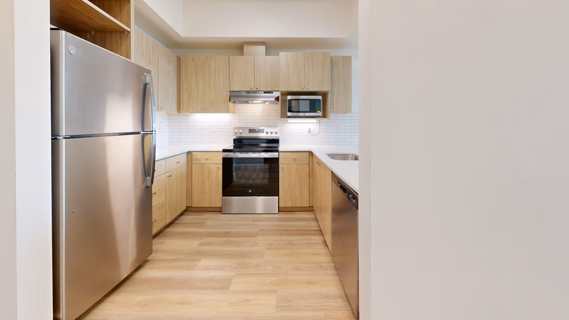 Modern kitchen with light wood cabinets, stainless steel refrigerator, oven, microwave, dishwasher, and white countertops with a white subway tile backsplash.