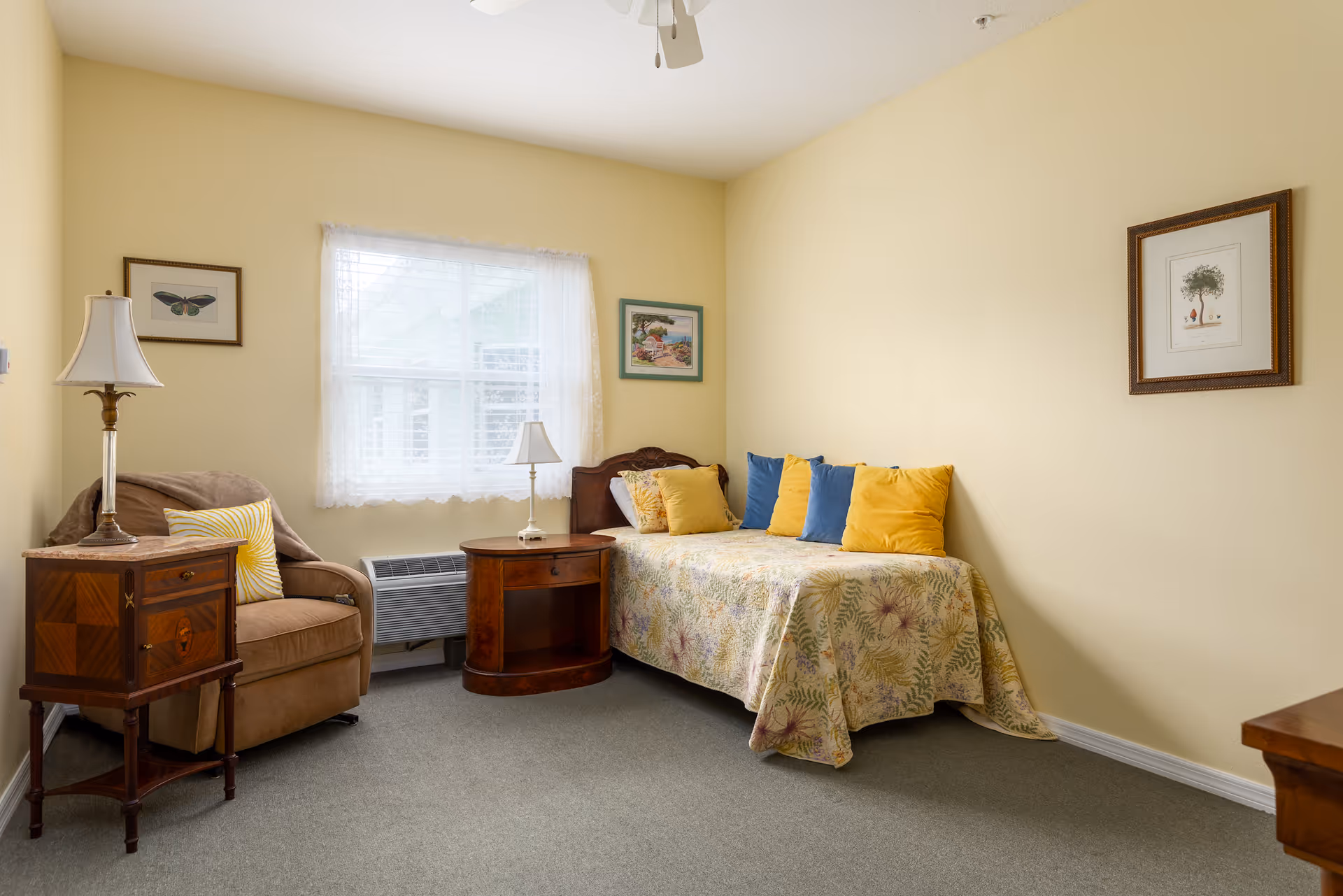 A cozy bedroom with a single bed covered in a floral quilt and decorated with yellow and blue pillows. Next to the bed is a wooden nightstand with a small lamp. There is a comfortable armchair with a yellow striped pillow and a wooden side table with a lamp beside it. The walls are painted light yellow and decorated with framed artwork. A window with sheer white curtains lets in natural light.