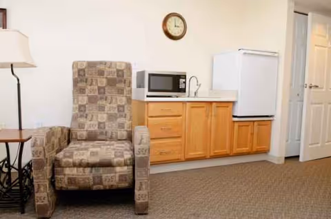 Cozy living area with a patterned armchair, microwave, mini fridge, and wooden cabinets along wall.
