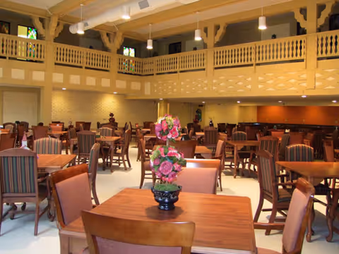 Spacious dining room with multiple wooden tables and chairs arranged neatly. A decorative flower arrangement is placed on the table in the foreground. The room features a high ceiling with exposed beams and a balcony railing along the upper level. Warm lighting illuminates the space, creating a welcoming atmosphere.