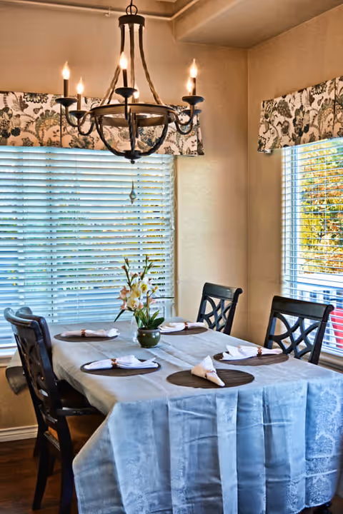 A dining room with a rectangular table covered with a light blue tablecloth. The table is set with six place settings, each with a round placemat and a folded white napkin. A small green vase with flowers is in the center of the table. Four dark wooden chairs with decorative backs surround the table. Two windows with white blinds and floral valances let in natural light. A black chandelier with six candle-style lights hangs above the table.