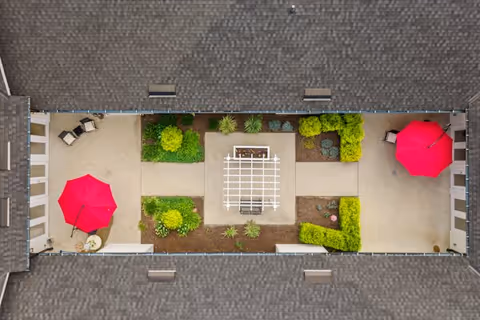 Aerial view of a rectangular courtyard enclosed by a building with a shingled roof. The courtyard features two bright red umbrellas shading seating areas on opposite ends, green shrubs and plants arranged symmetrically along the sides, and a white pergola structure in the center with a bench underneath.