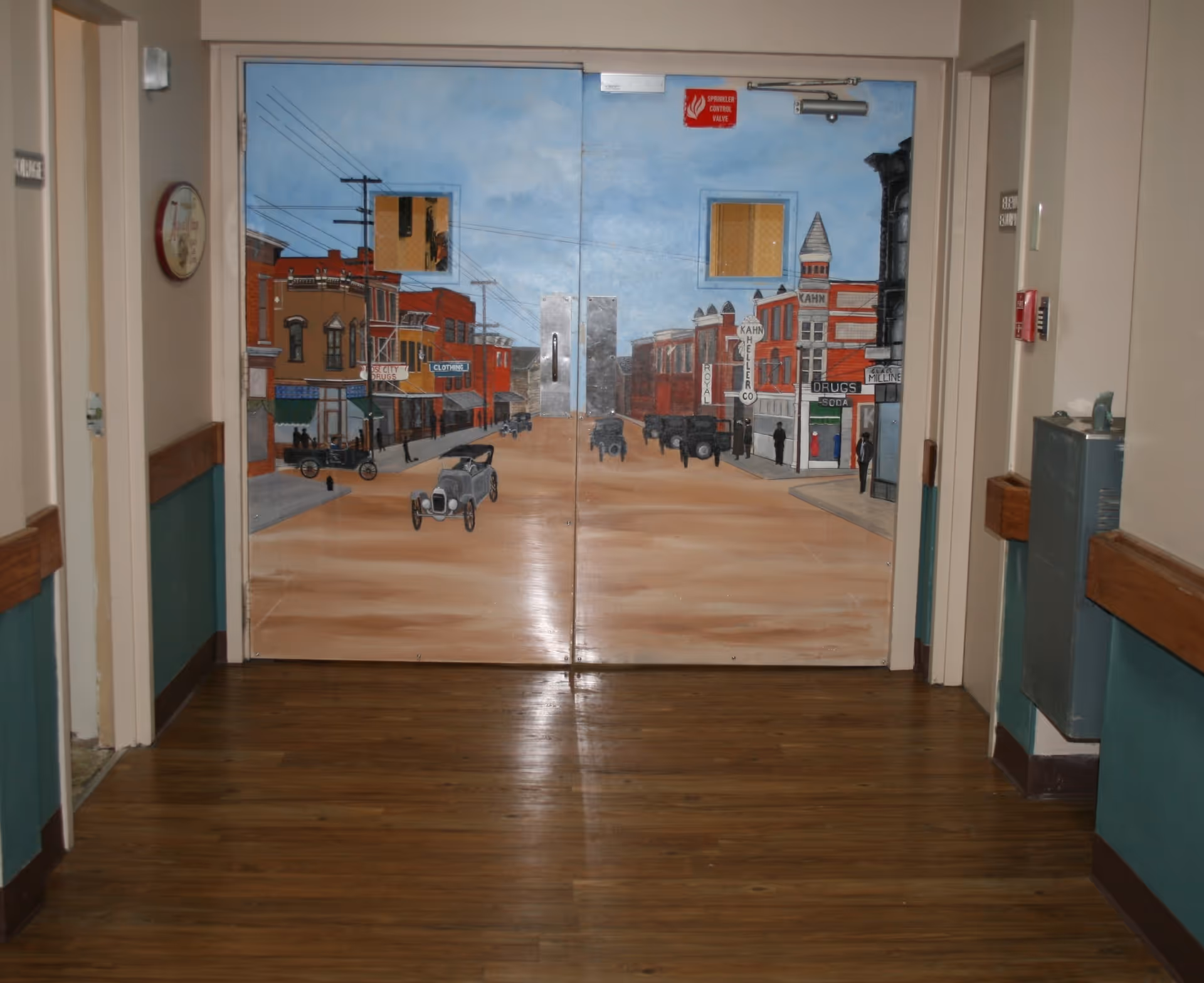 Double doors at the end of a hallway painted with a mural depicting a vintage street scene with old cars, buildings, and people. The hallway has wooden floors, teal and beige walls with wooden handrails, and a clock on the left wall.