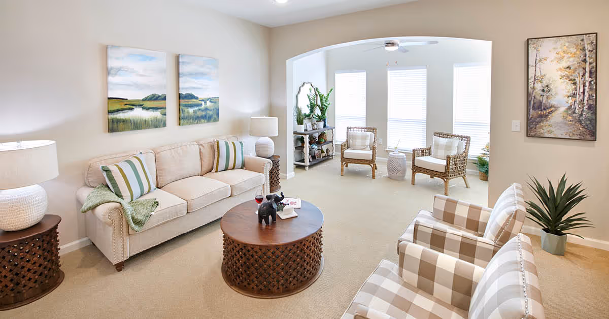 A bright and cozy living room with a beige sofa adorned with striped pillows and a green throw blanket. There are two plaid armchairs, a round wooden coffee table with decorative items, and two wicker chairs near large windows letting in natural light. The room is decorated with landscape paintings, a potted plant, and a side table with a lamp.