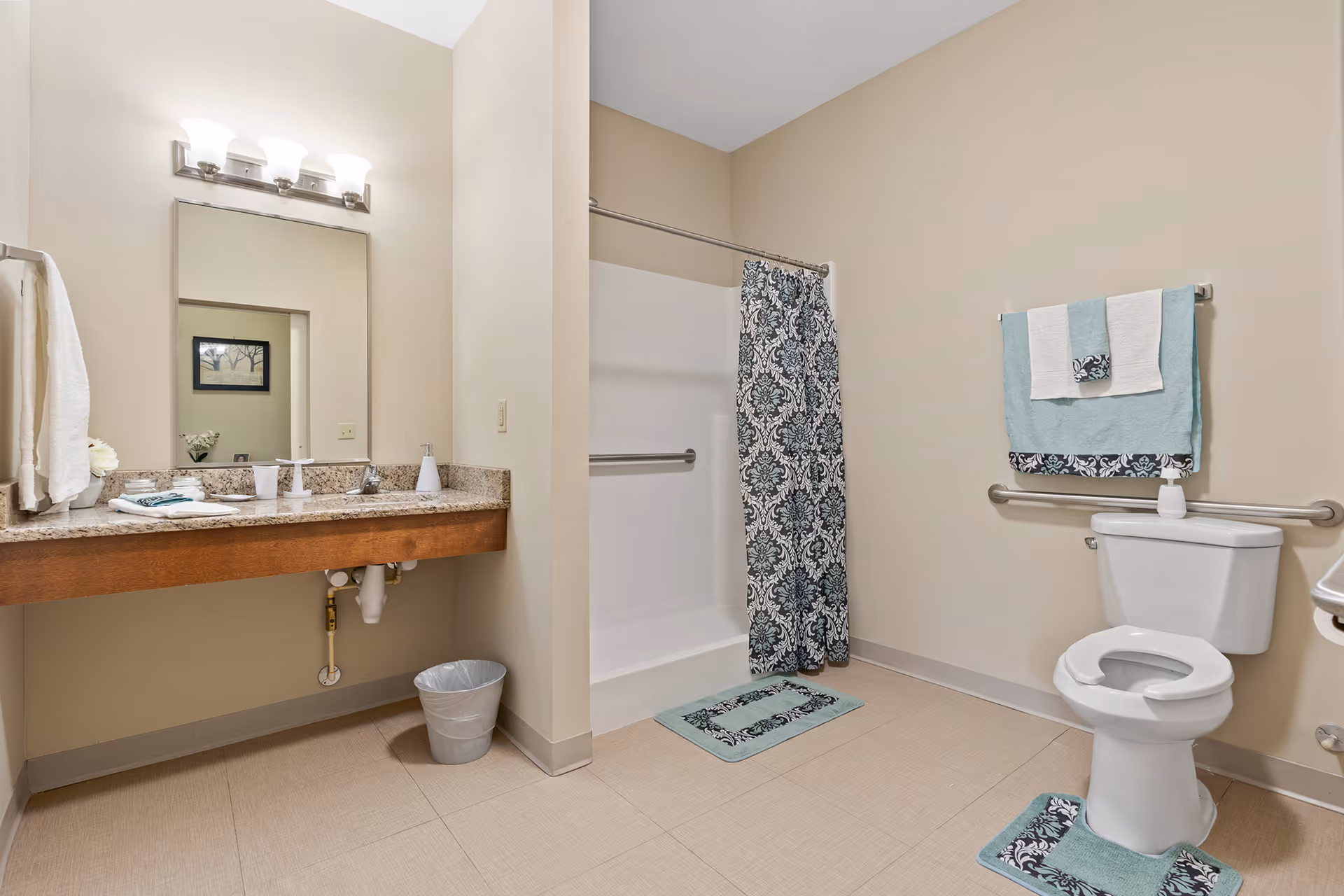 A clean and spacious bathroom with beige walls and tiled floor. It features a white toilet with a grab bar behind it, a shower with a patterned curtain, and a long granite countertop with a sink, mirror, and light fixture above. Towels and bath mats in teal and black patterns are neatly arranged.