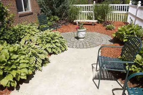 Outdoor courtyard with circular stone centerpiece, green chairs, bench, and surrounding shrubs.