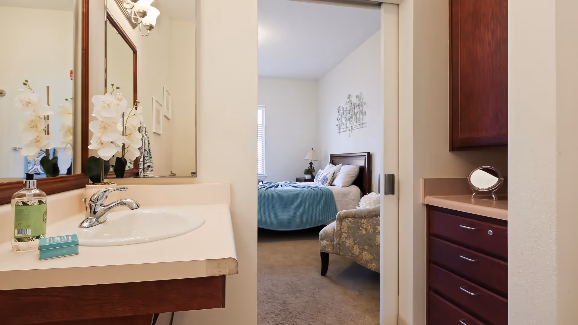 View of a bedroom through an open doorway from a bathroom area. The bathroom has a sink with a faucet, a bottle of hand soap, and a small box on the counter. The bedroom features a bed with a blue blanket, pillows, a bedside table with a lamp, and a floral armchair. There is a decorative wall piece above the bed and a window letting in natural light.