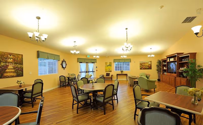 A spacious, well-lit common area with multiple round tables and chairs arranged for seating. The room features wooden flooring, several chandeliers hanging from the ceiling, large windows with blinds and valances, and framed artwork on the walls. There is a wooden entertainment center with a television and decorative plants, as well as comfortable armchairs near the back of the room.