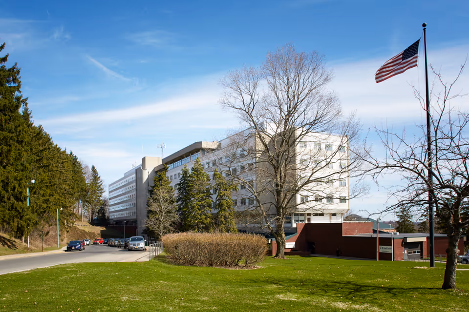 Front exterior of a multi-story rehabilitation and nursing facility with trees, lawn, parked cars, and an American flag.