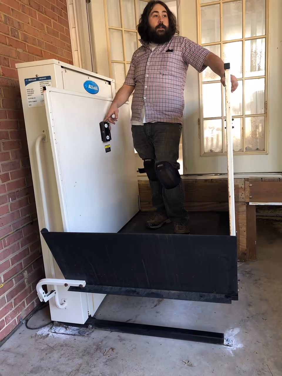 A man with knee pads stands on a wheelchair lift platform outside a building entrance with glass-paneled doors. The lift is attached to a brick wall and has a white control panel with a blue 'Access' label.