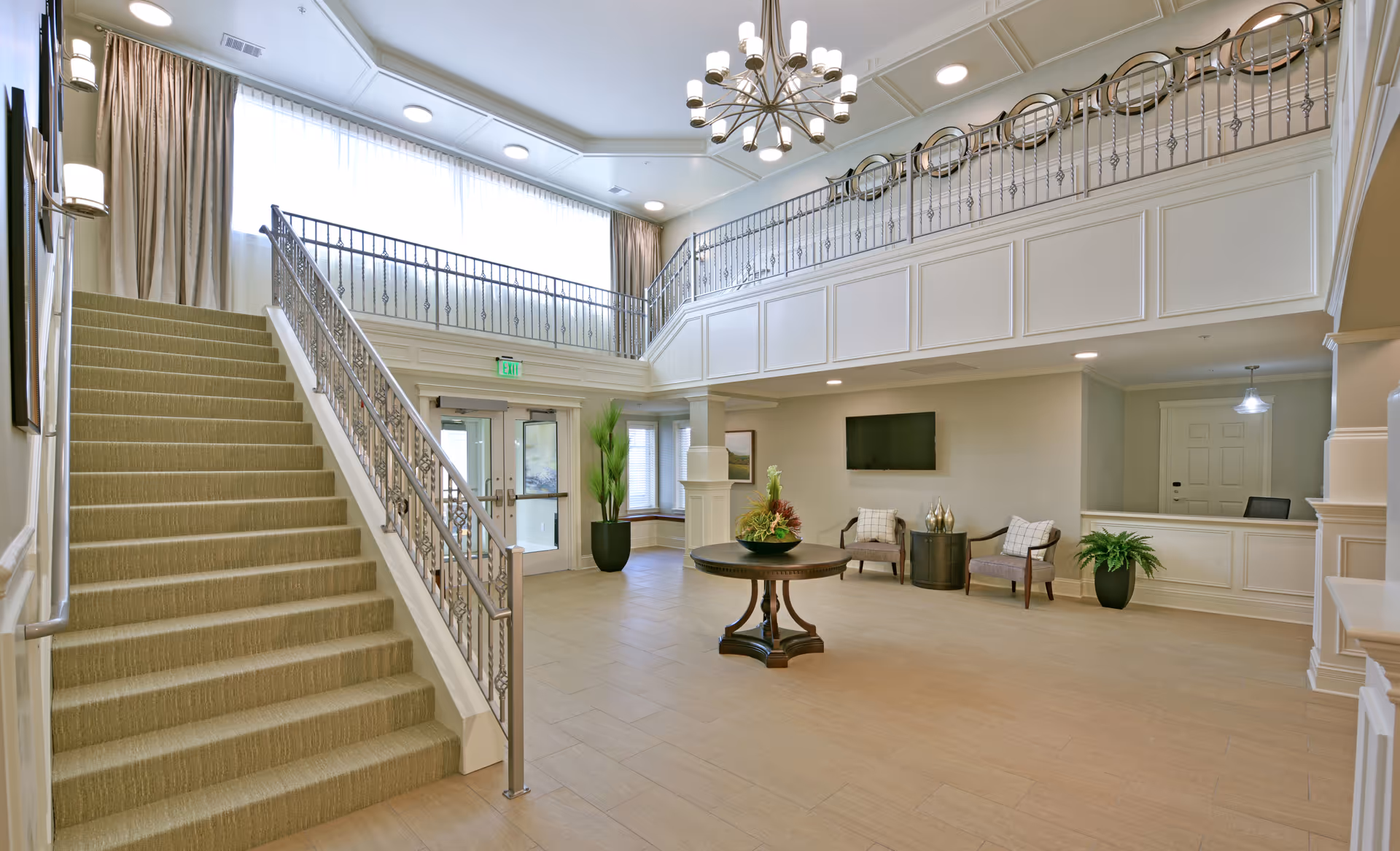 Spacious and well-lit senior living facility lobby with a carpeted staircase on the left, a round wooden table with a floral arrangement in the center, two armchairs with cushions, a wall-mounted TV, potted plants, and a reception desk on the right. The area features large windows with curtains, a chandelier, and a balcony railing above.