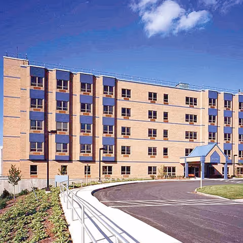 Exterior view of a multi-story brick building with blue accents around the windows and a blue entrance canopy. The building is surrounded by a paved driveway, a curved sidewalk with a railing, and landscaped greenery under a partly cloudy sky.
