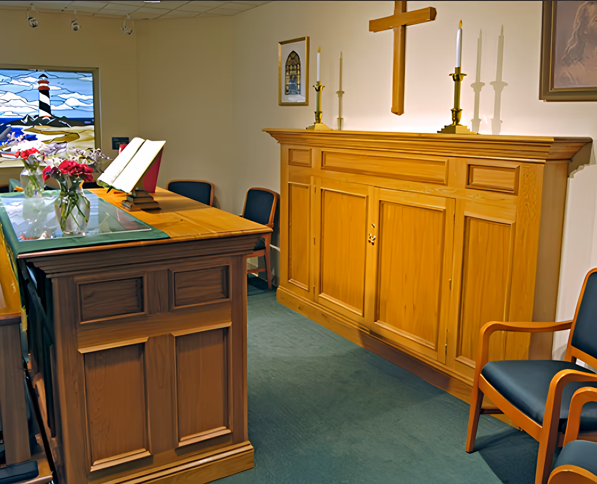 Small chapel interior with a wooden altar and lectern, wall cross, candles, stained glass window and chairs.