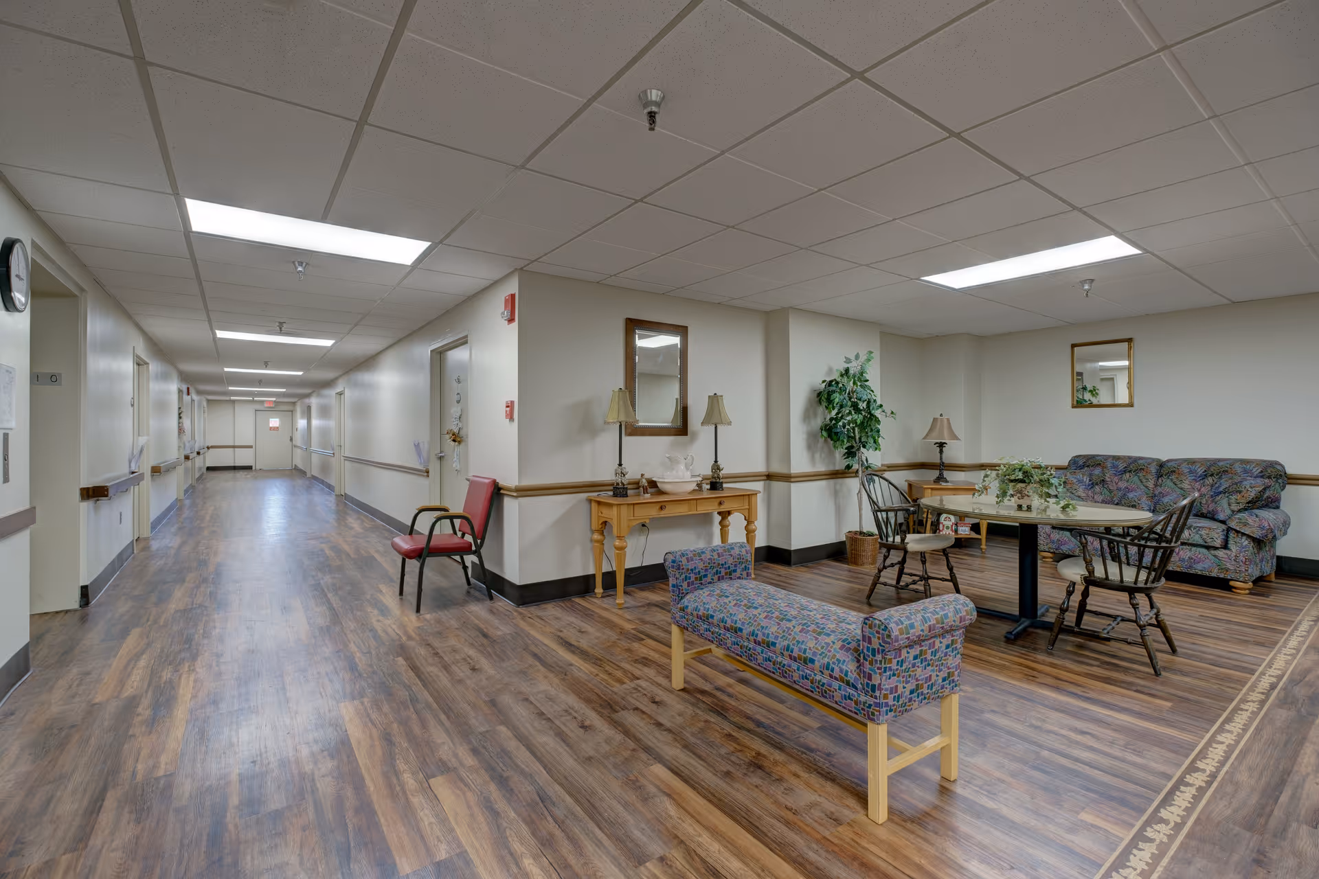 A long hallway in a senior living facility with wood flooring and white walls. On the right side, there is a sitting area with a patterned bench, a table with two chairs, a floral couch, a wooden side table with a lamp, a small wooden console table with two lamps and a mirror above it, and a potted plant. The ceiling has fluorescent lighting panels.