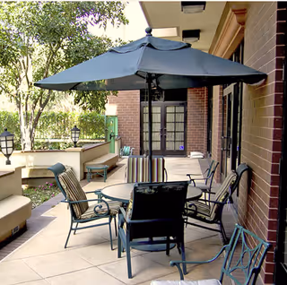 Outdoor patio with a round table, large umbrella and several chairs beside a brick building and garden.