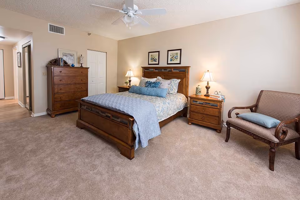 A well-lit bedroom with a wooden bed frame, a bed made with patterned bedding and multiple pillows, two wooden nightstands with lamps, a wooden dresser with books and decor, a cushioned wooden armchair with a pillow, beige carpet, and a ceiling fan.