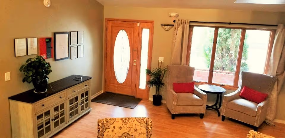 A cozy living room area with two beige armchairs each with a red pillow, a small round black table between them, a large window with beige curtains, a wooden front door with an oval glass panel, a wooden cabinet with glass doors topped with a potted plant, and framed documents or artwork on the wall.