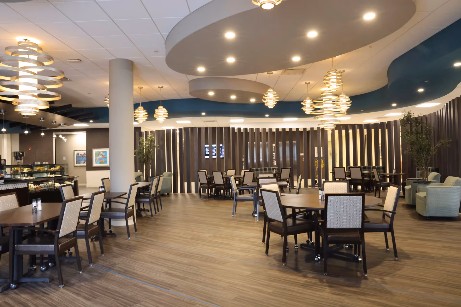 A spacious and modern dining area with multiple round and rectangular tables surrounded by chairs. The ceiling features circular recessed lighting and decorative hanging light fixtures. The floor is wood-patterned, and there are some plants and artwork on the walls. A food display counter is visible on the left side of the room.