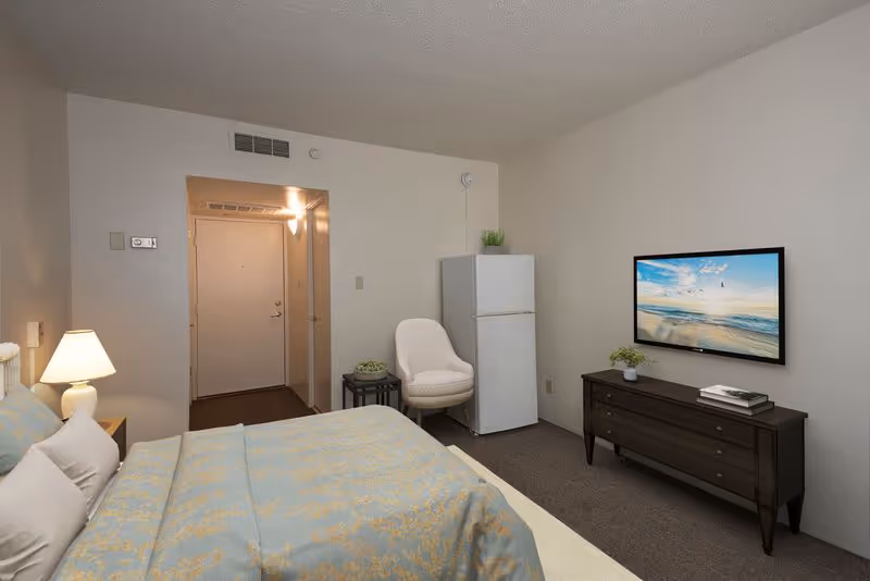 A furnished studio bedroom with a bed, nightstand and lamp, armchair, small refrigerator, dresser with a wall-mounted TV, and an entryway.