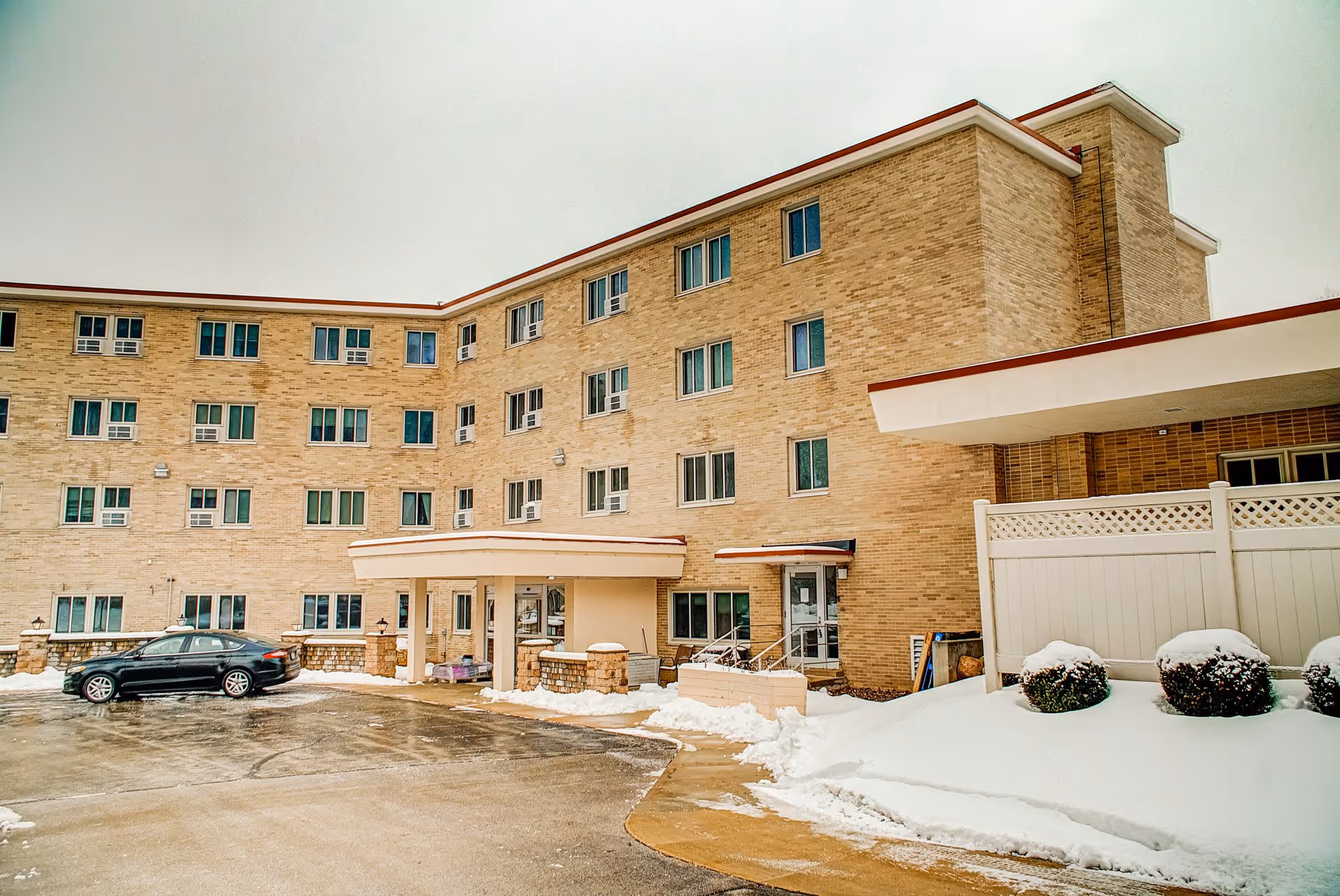 Front exterior of a multi-story brick building with a covered entrance, a parked car, and snow on the ground.